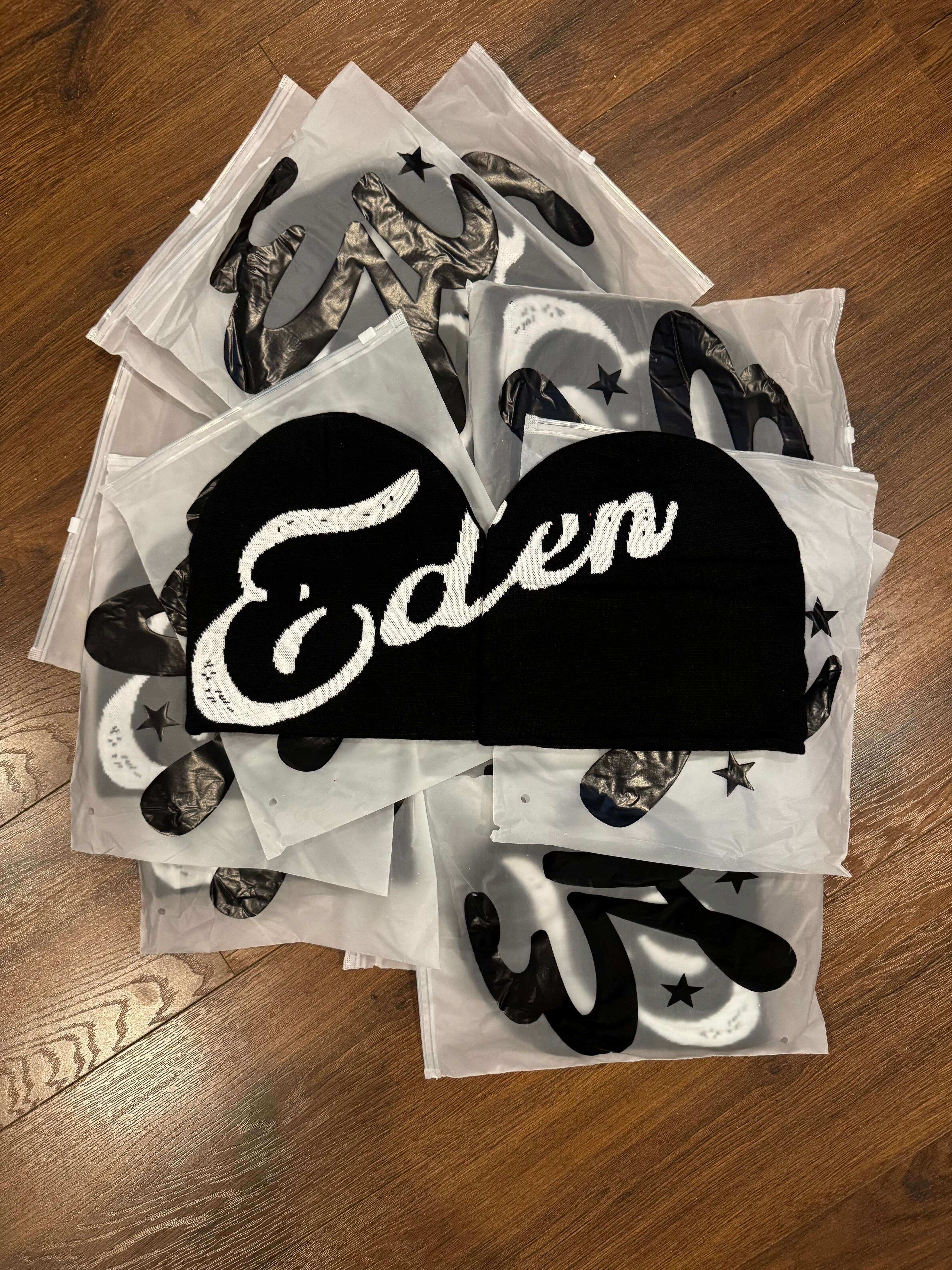 EDEN Cursive Beanie displayed among various other hats in packaging on a wooden surface