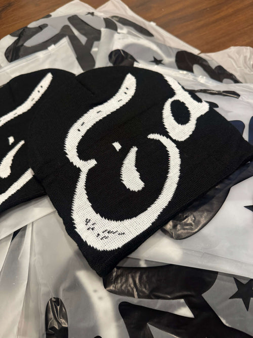 EDEN Cursive Beanie displayed on a wooden surface with patterned bags in the background.